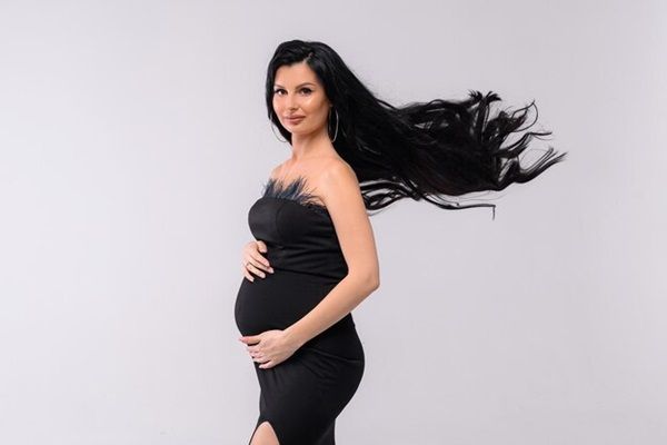 bella femmina che abbraccia il ventre incinta con trucco professionale bacchetta capelli lunghi ondulati indossando in abito da sera elegante mentre posa in interni