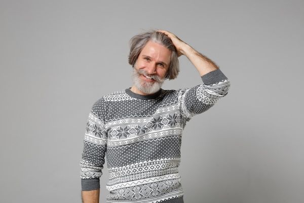 Anziano sorridente con capelli grigi, baffi e barba, uomo in maglione di lana, posa isolata su sfondo grigio, ritratto in studio. Concetto di stile di vita delle persone. Spazio per il testo. Si mette la mano sulla testa.