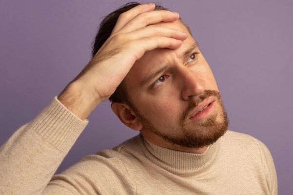 Uomo che indossa un maglione beige dal taglio alto su sfondo viola che guarda di lato con un'espressione dubbiosa.