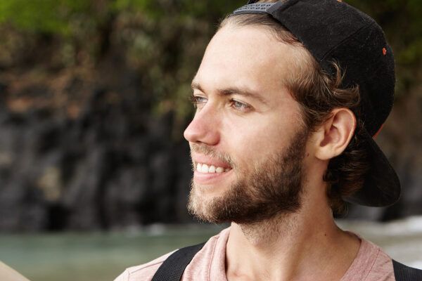 Giovane con gli occhi chiari e la barba che indossa un berretto sulla spiaggia guardando l'orizzonte.
