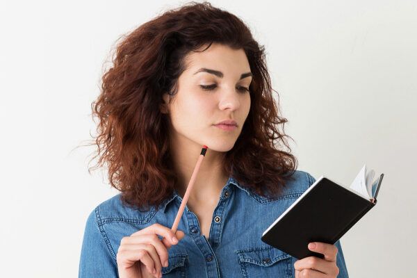 Donna con camicia di jeans blu e bottoni, capelli asciutti e ondulati che guarda l'agenda in mano mentre tiene la matita con l'altra - Riparare i capelli danneggiati