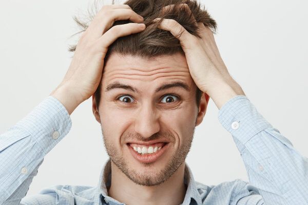 Giovane con capelli castani e pelle chiara con un'espressione preoccupata mentre si strappa i capelli dalle radici con entrambe le mani - La forfora negli uomini.