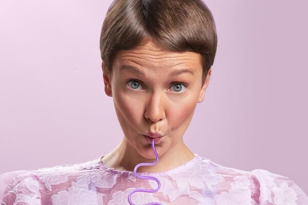 Donna con i capelli corti e gli occhi chiari in posa su uno sfondo color pastello mentre sorseggia una sigaretta con un'espressione particolare.