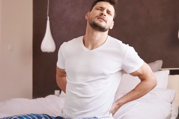 Giovane con la barba in pigiama di flanella bianca che tiene la parte bassa della schiena mentre si alza dal letto.