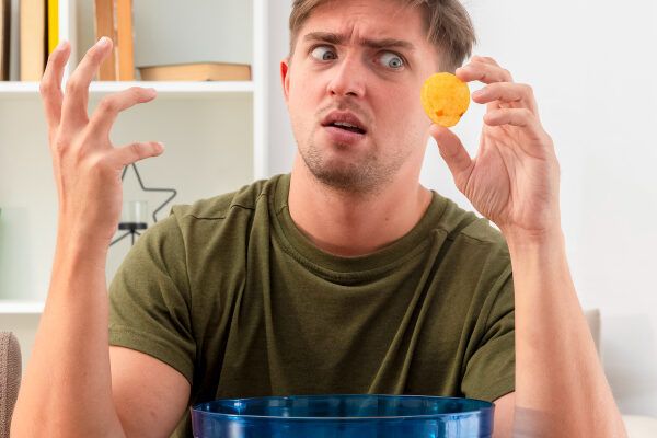 Uomo biondo con gli occhi chiari con un'espressione confusa mentre guarda una patatina fritta che ha tra le dita.
