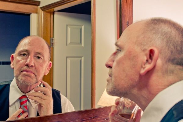 un uomo che si guarda allo specchio e si mette il profumo, presenta segni di alopecia