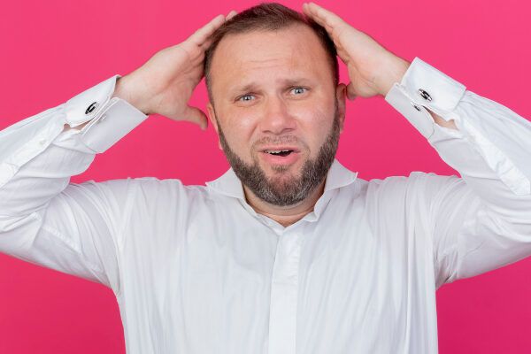 Uomo in camicia bianca che soffre di alopecia in posa su uno sfondo rosa guardando la telecamera con un'espressione di frustrazione e preoccupazione.