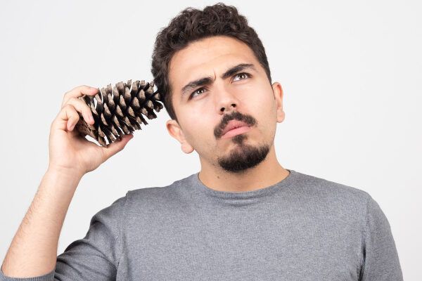 Uomo in maglione grigio con barba e baffi che alza lo sguardo pensieroso mentre tiene un ananas in mano e lo appoggia sulla testa.