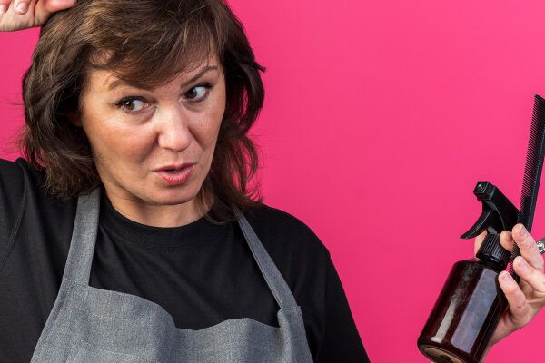 Donna parrucchiera con strumenti per capelli in mano mentre prende una ciocca di capelli mentre l'altra guarda di lato in modo sorpreso.