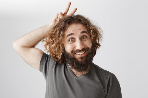 Uomo di origine mediorientale con i capelli lunghi che sorride alla telecamera con un'espressione divertente sulle mani dietro la testa - Curiosità sui capelli degli uomini.