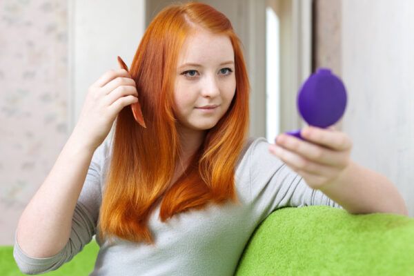 Ragazza dai capelli rossi che si guarda allo specchio in mano mentre sorride e si pettina delicatamente.