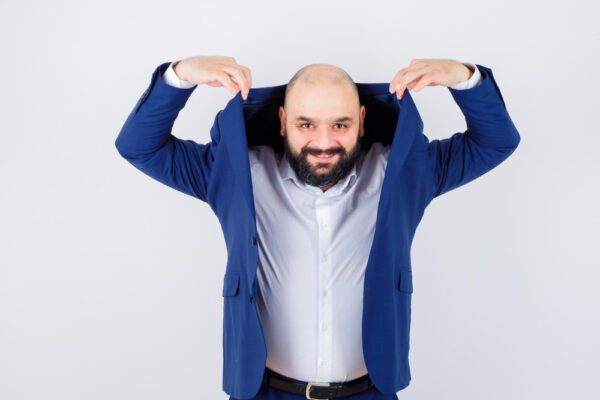 Uomo con alopecia areata e giacca blu che la solleva sulle spalle mentre guarda la telecamera.