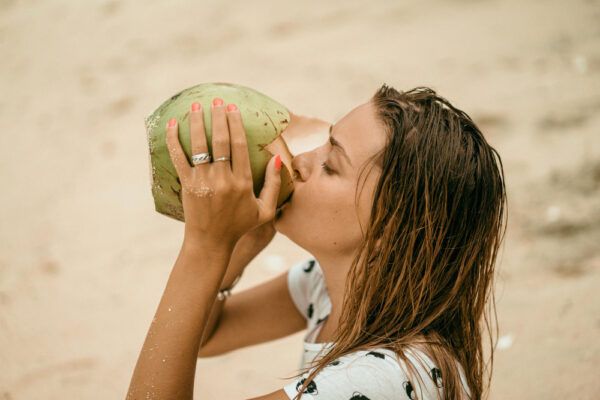Ragazza dai capelli unti che beve acqua da una noce di cocco sulla riva della spiaggia - Olio di cocco per capelli.