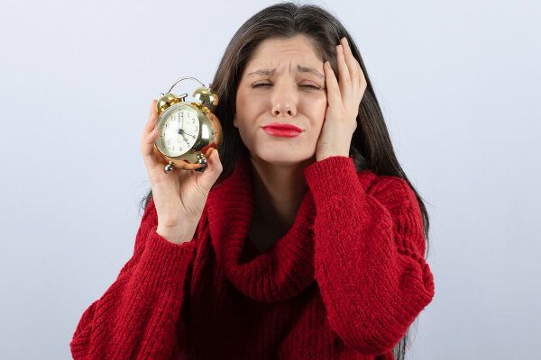 Donna con capelli castano scuro in un maglione e labbra rosse che ascolta un orologio tra le mani.