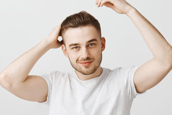 Giovane con gli occhi chiari, la pelle illuminata e una bella barba che sorride alla telecamera mentre si accarezza i capelli - Capelli sani dall'interno.