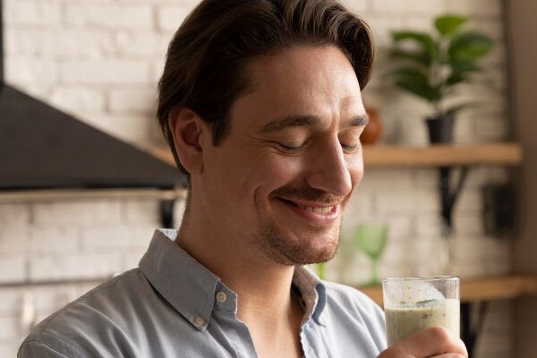 Uomo con i capelli castani in cucina con in mano un drink in una tazza di vetro.