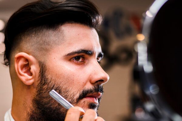 Giovane che si guarda allo specchio mentre si taglia la barba con un rasoio.