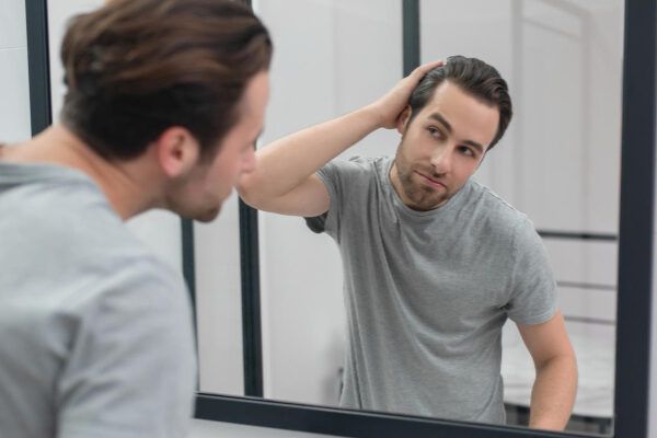 Bel giovane che studia i suoi capelli mentre guarda il suo riflesso nello specchio del bagno - Fermare l´alopecia giovanile.