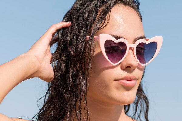 Giovane donna con capelli ondulati che indossa occhiali da sole rosa a forma di cuore in posa mentre prende il sole.