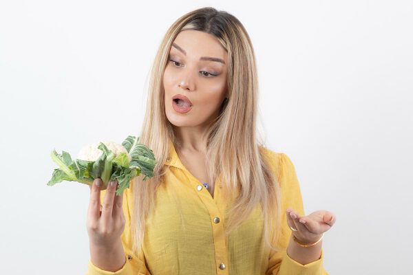 Donna bionda con i capelli lunghi e una camicetta gialla che guarda con espressione sorpresa un cavolfiore che ha tra le mani - Vegetariani fa perdere i capelli.