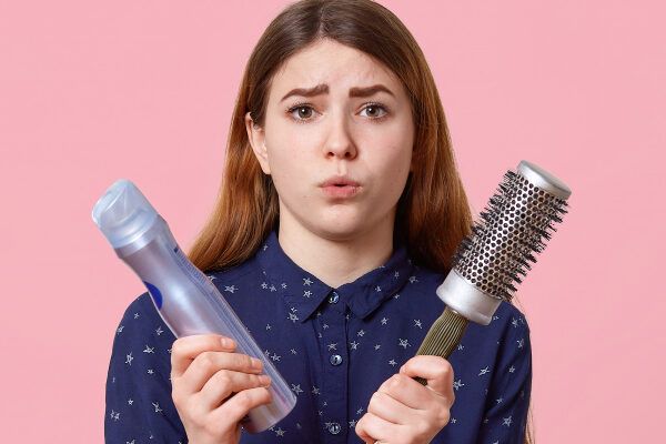 Donna che tiene un prodotto in mano e una spazzola rotonda per lo styling nell'altra con aria spaventata - Prodotti per fissare i capelli.