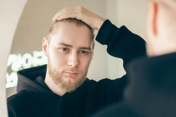 Un uomo biondo con la barba, la pelle chiara e una felpa nera che si guarda allo specchio mentre si accarezza i capelli - Sindrome dei capelli impettinabili.