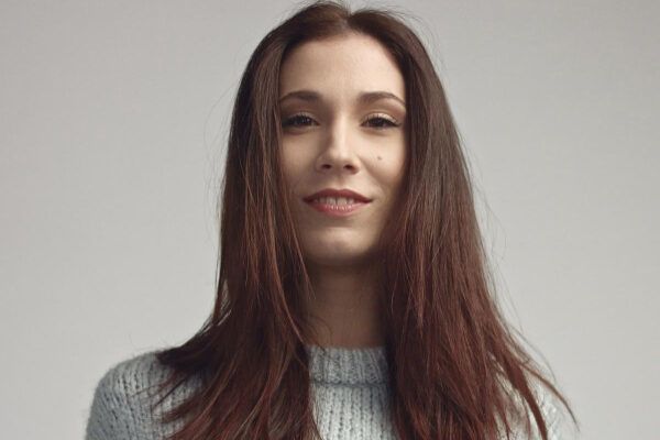 Donna con maglione grigio e capelli lunghi fino alle spalle che sorride alla telecamera con uno sguardo spettinato.