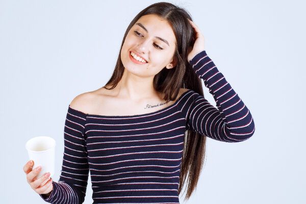Giovane ragazza con i capelli lisci e un maglione a righe che sorride di lato con un bicchiere in mano.