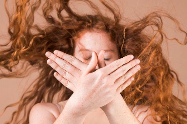 Donna dai capelli rossi con riccioli arruffati e fuori posto che le coprono il viso con entrambe le mani puntate verso la telecamera.