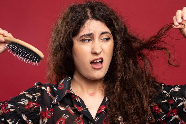 Donna con capelli ondulati in posa su uno sfondo bordeaux mentre guarda di lato con un'espressione disgustata e una spazzola cellulare in mano.