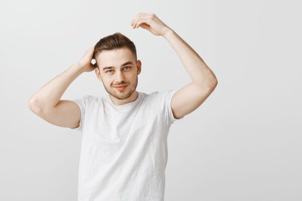 Giovane ragazzo in flanella bianca in posa su sfondo bianco mentre guarda la telecamera sorridente mentre si tocca i capelli con entrambe le mani.