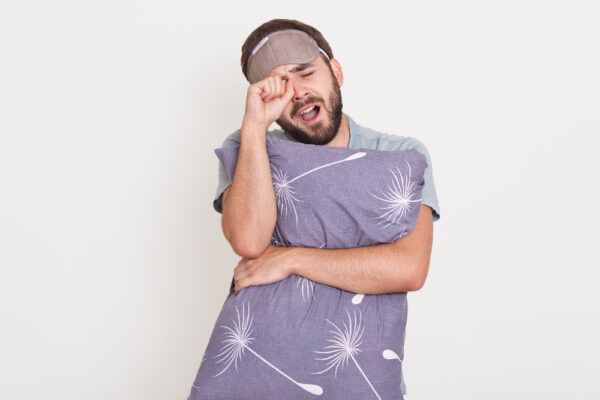 Ragazzo che sbadiglia con la maschera per dormire, abbraccia un cuscino e si stropiccia gli occhi.