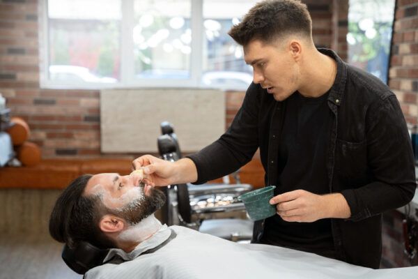 Barbiere nel suo spazio di lavoro mentre applica la maschera al suo cliente che ha la barba lunga.