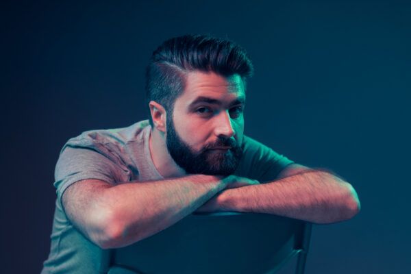 Uomo con taglio di capelli lungo e barba seduto su una sedia in posa per la fotocamera.