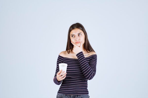 Giovane ragazza con lunghi capelli lisci che si tocca il mento con le dita in espressione dua mentre tiene un bicchiere con l'altra mano.