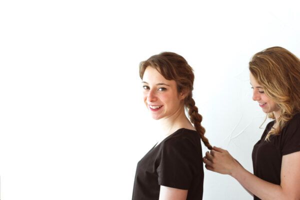 Una ragazza bionda con i capelli mossi pettina i capelli di un'altra donna creando una treccia mentre sorride alla telecamera.