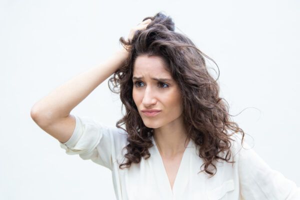 Una ragazza con i capelli ricci si gratta la testa con un'espressione lamentosa mentre guarda di lato.
