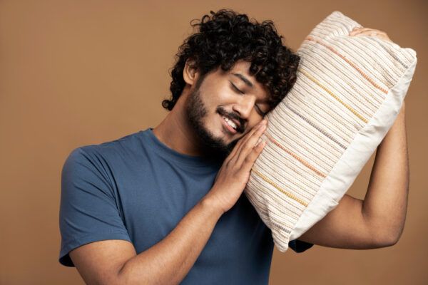Ragazzo dalla pelle scura, con i capelli ricci e la barba, che sorride mentre è sdraiato su un cuscino.