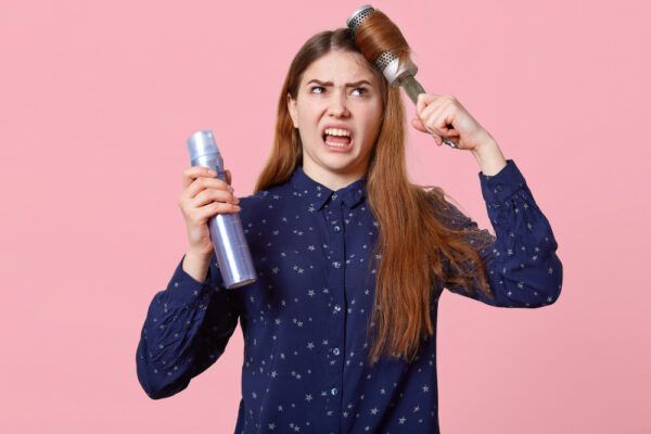 Una ragazzina si pettina i capelli con una spazzola rotonda e tiene in mano una sostanza chimica, mentre la guarda con un'espressione lamentosa.
