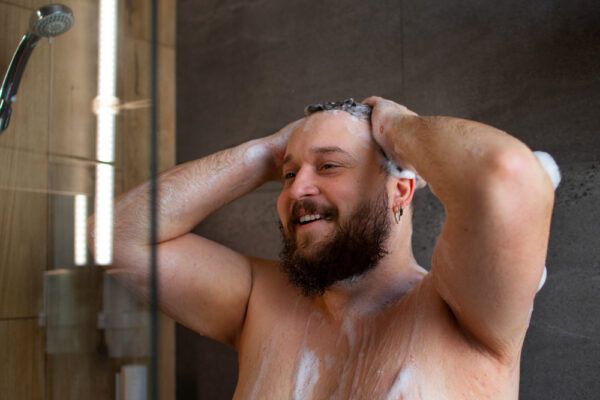 Uomo con barba e capelli castano scuro e molta schiuma che si lava i capelli sotto la doccia - Shampoo dopo il trapianto.