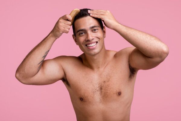 Bellissimo ragazzo, ben muscoloso e con gli occhi chiari che si pettina i capelli mentre sorride alla telecamera e posa su uno sfondo rosa.