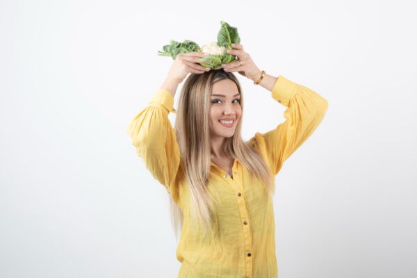 Ragazza bionda su sfondo bianco con maglione giallo in posa con un cavolfiore in testa.