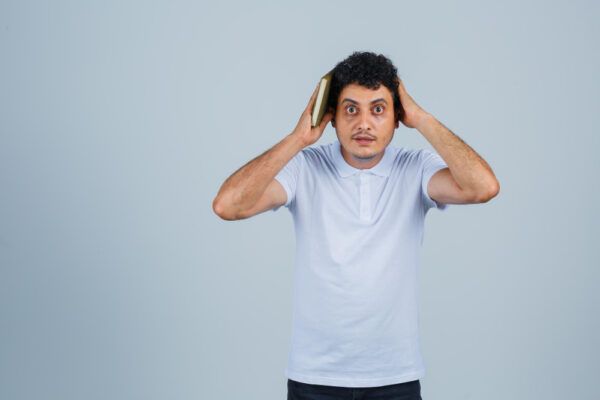 Un ragazzo con occhiaie e problemi di sonno si tocca la testa per la sorpresa mentre guarda la telecamera e tiene in mano un libro.