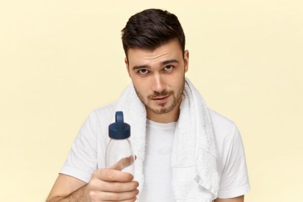 Un giovane sportivo con un asciugamano al collo, mentre tiene in mano un thermos e guarda la telecamera in modo seducente.