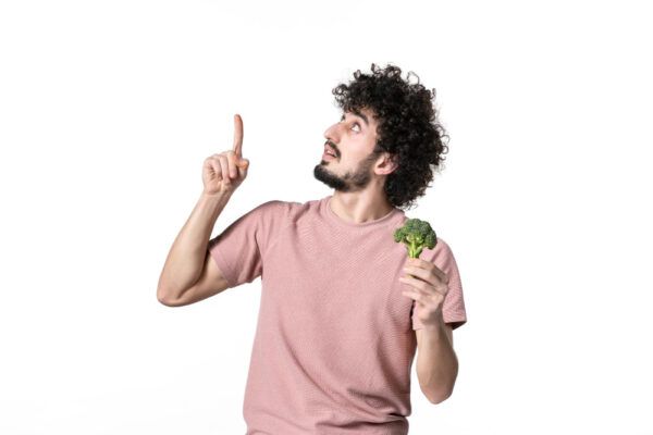 Un uomo dai capelli ricci in posa su uno sfondo bianco indicando verso l'alto mentre tiene in mano un broccolo.