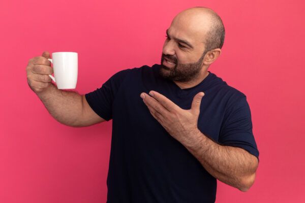 Uomo con alopecia in posa su uno sfondo rosa mentre guarda dubbioso e indica una tazza di caffè.