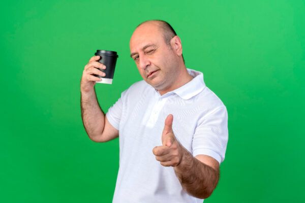 Uomo adulto con alopecia che mostra un bicchiere di caffè mentre posa davanti alla telecamera con un occhiolino.